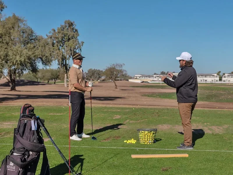 Two men stand on a grassy driving range with golf clubs, one instructing the other. A bucket of golf balls and golf bag are nearby. Trees and buildings are visible in the background under a clear blue sky.