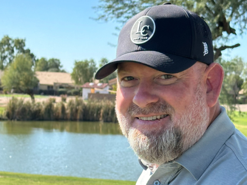 A bearded man in a gray polo shirt and black cap smiles outdoors near a pond, with green grass and trees under a clear blue sky in the background.